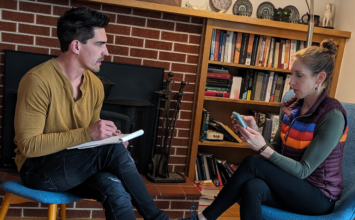 A man and woman are engaged in a discussion, with the woman looking at her phone while the man takes notes. The setting features a living room with a brick wall and a bookshelf in the background.