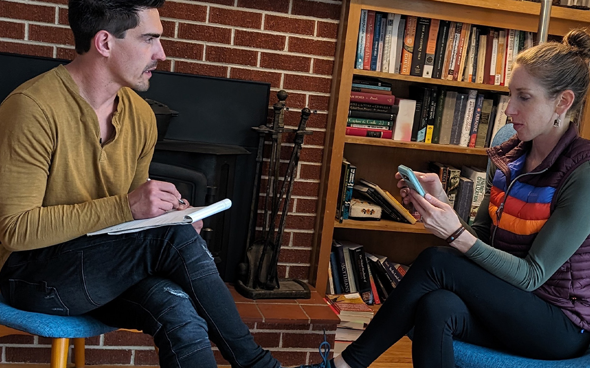 A man and woman are engaged in a discussion, with the woman looking at her phone while the man takes notes. The setting features a living room with a brick wall and a bookshelf in the background.