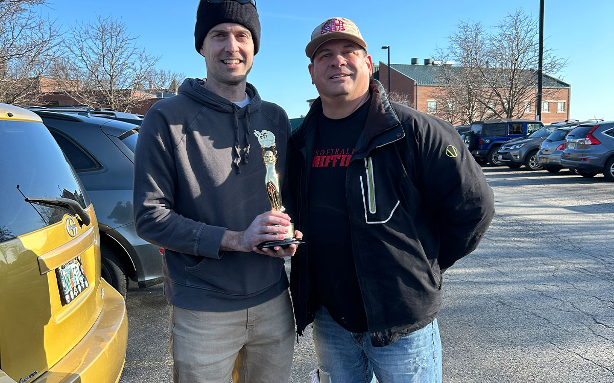 David Safin and Don Hartman stand together in a parking lot, one holding a trophy.