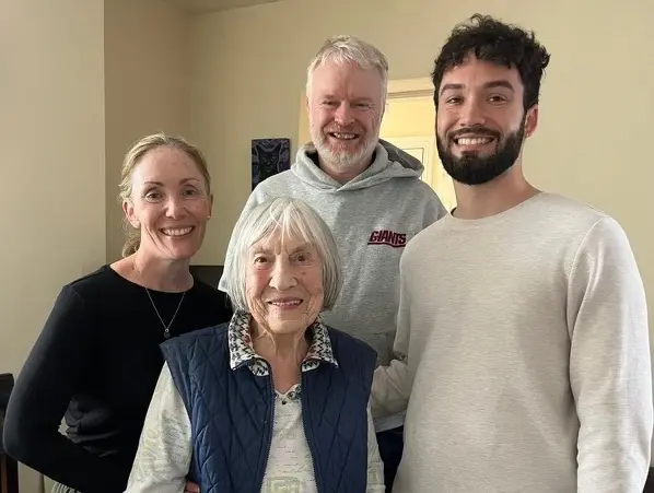 A group of four family members posing together, including an elderly woman in a blue vest and three younger adults, all smiling warmly.