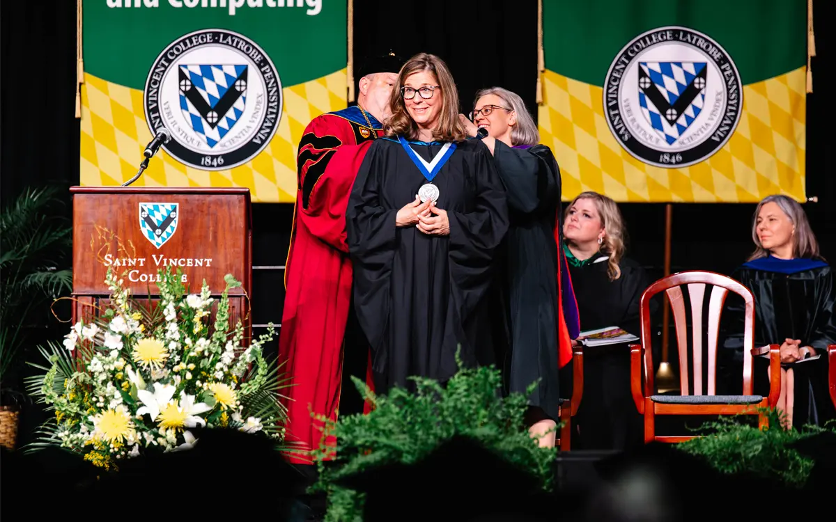 A faculty member receiving an award on stage during a graduation ceremony at Saint Vincent College, with colorful banners in the background.