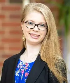 A professional headshot of a woman with long blonde hair and glasses, wearing a black blazer over a floral blue blouse, set against a brick background.