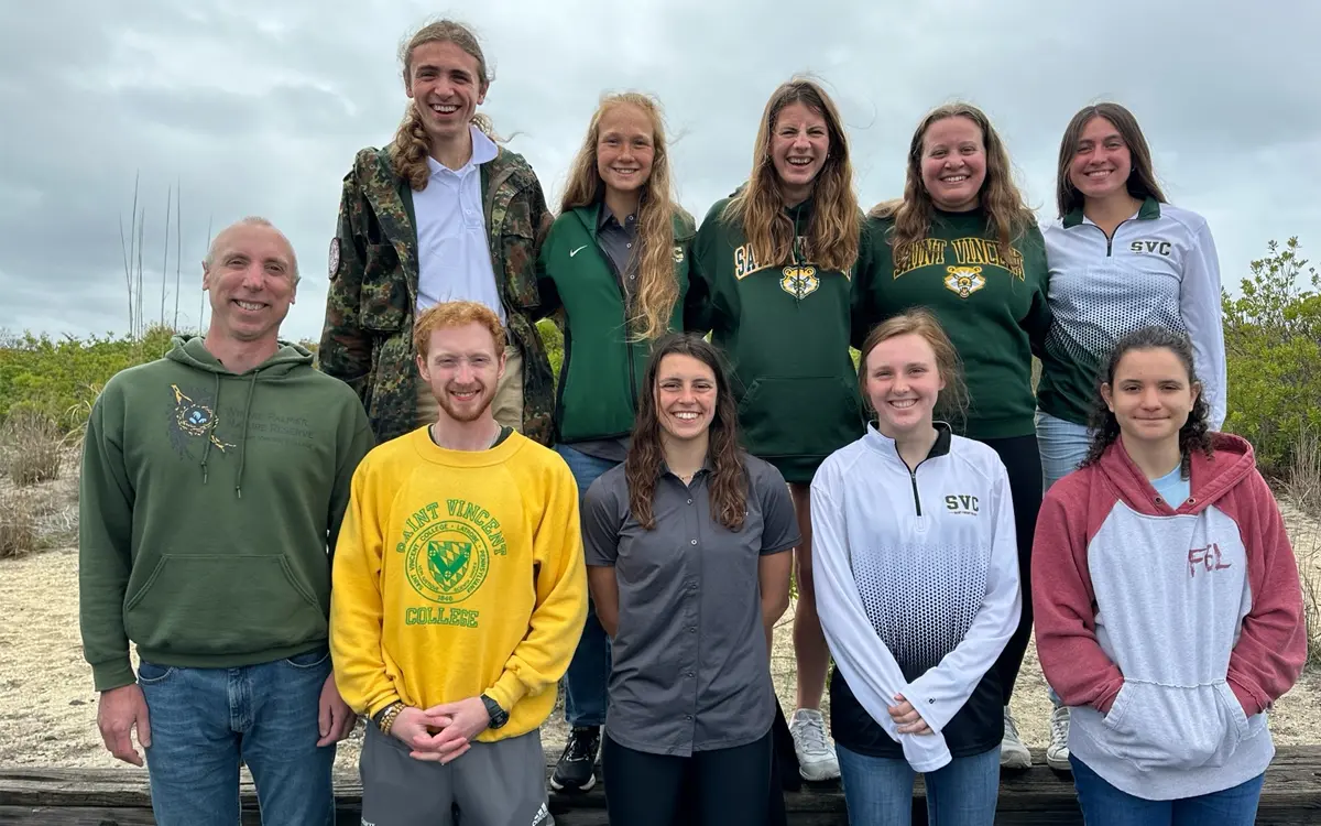 A group photo of twelve students and faculty members, smiling and posing outdoors, wearing casual college-branded attire.