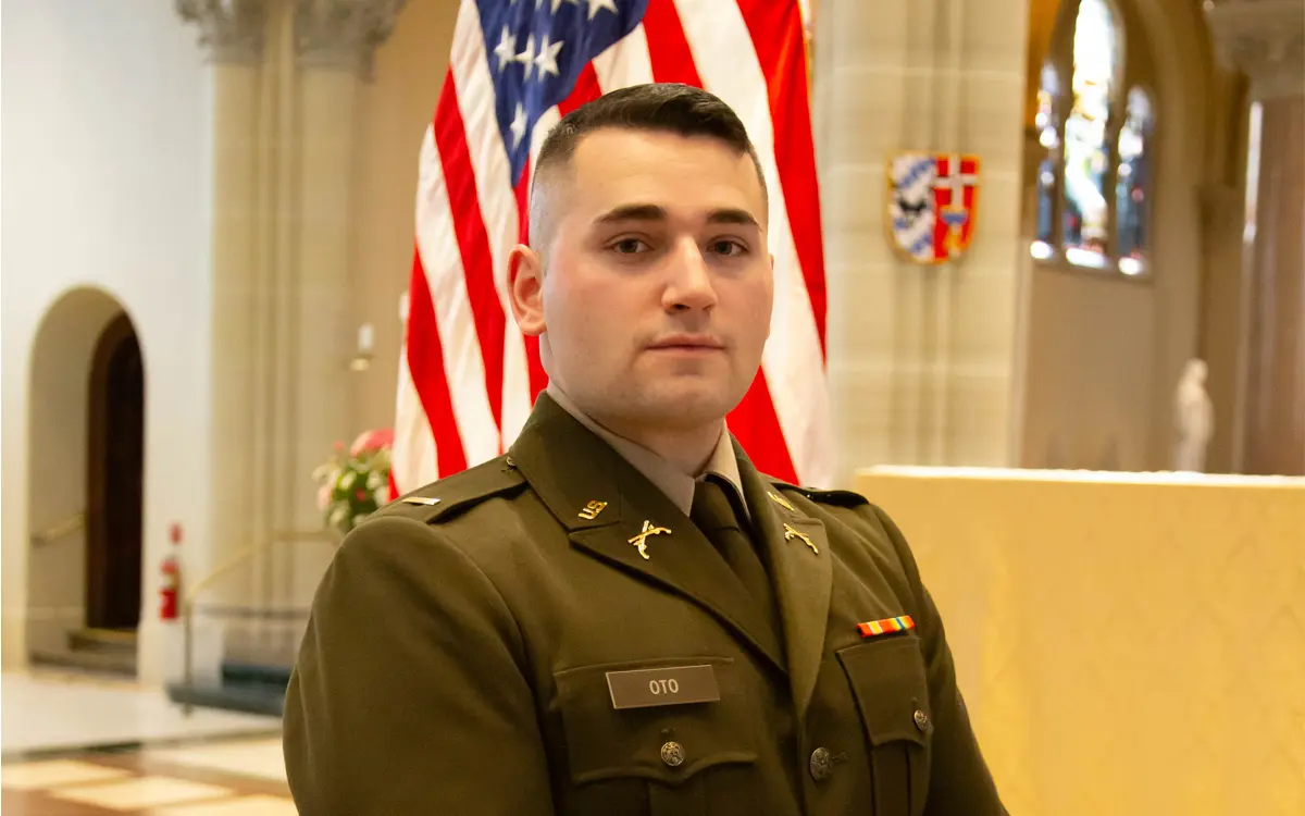 A soldier in uniform poses in front of an American flag, inside a beautifully lit venue.