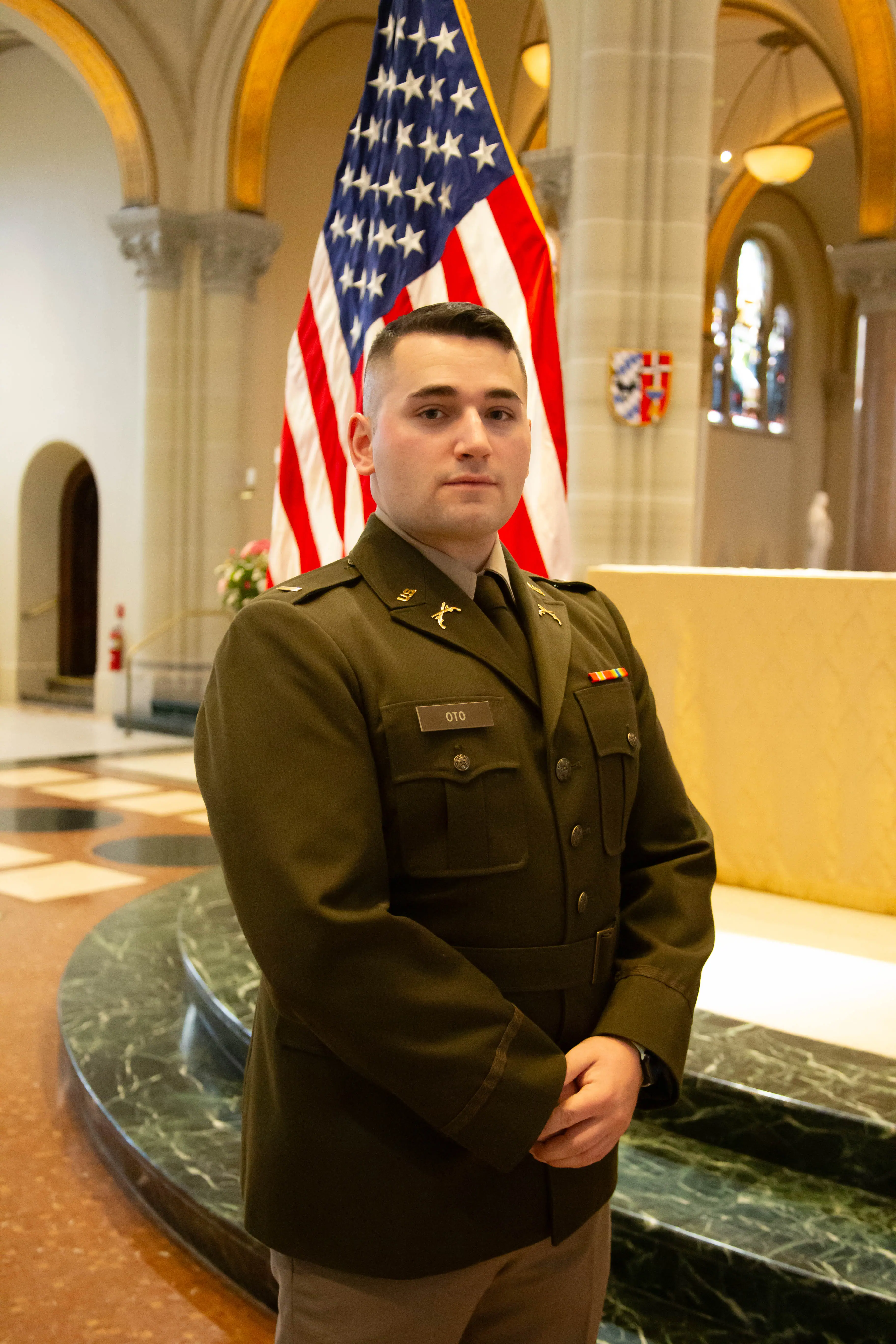 A soldier in uniform poses in front of an American flag, inside a beautifully lit venue.