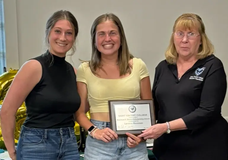 Three women at an awards ceremony, with one holding a certificate for an achievement while the others stand beside her, smiling.