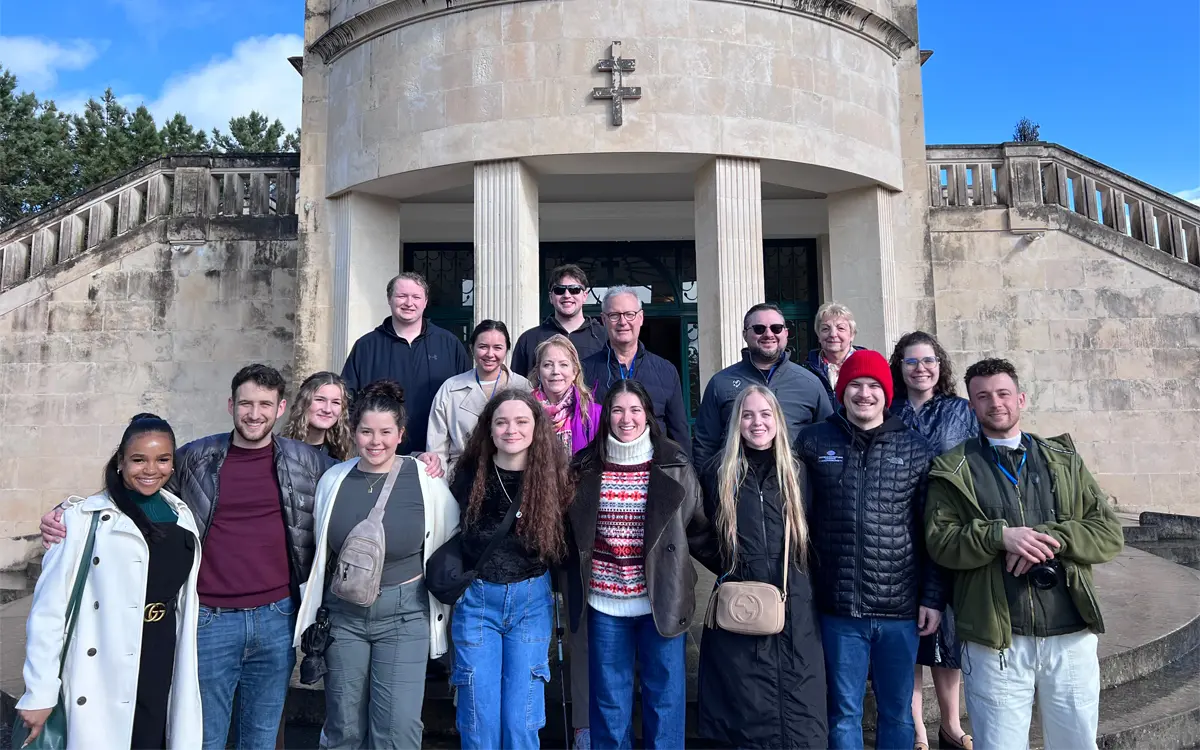 A group photo of 20 people gathered outside a building with a cross symbol, showcasing a diverse mix of individuals smiling together.