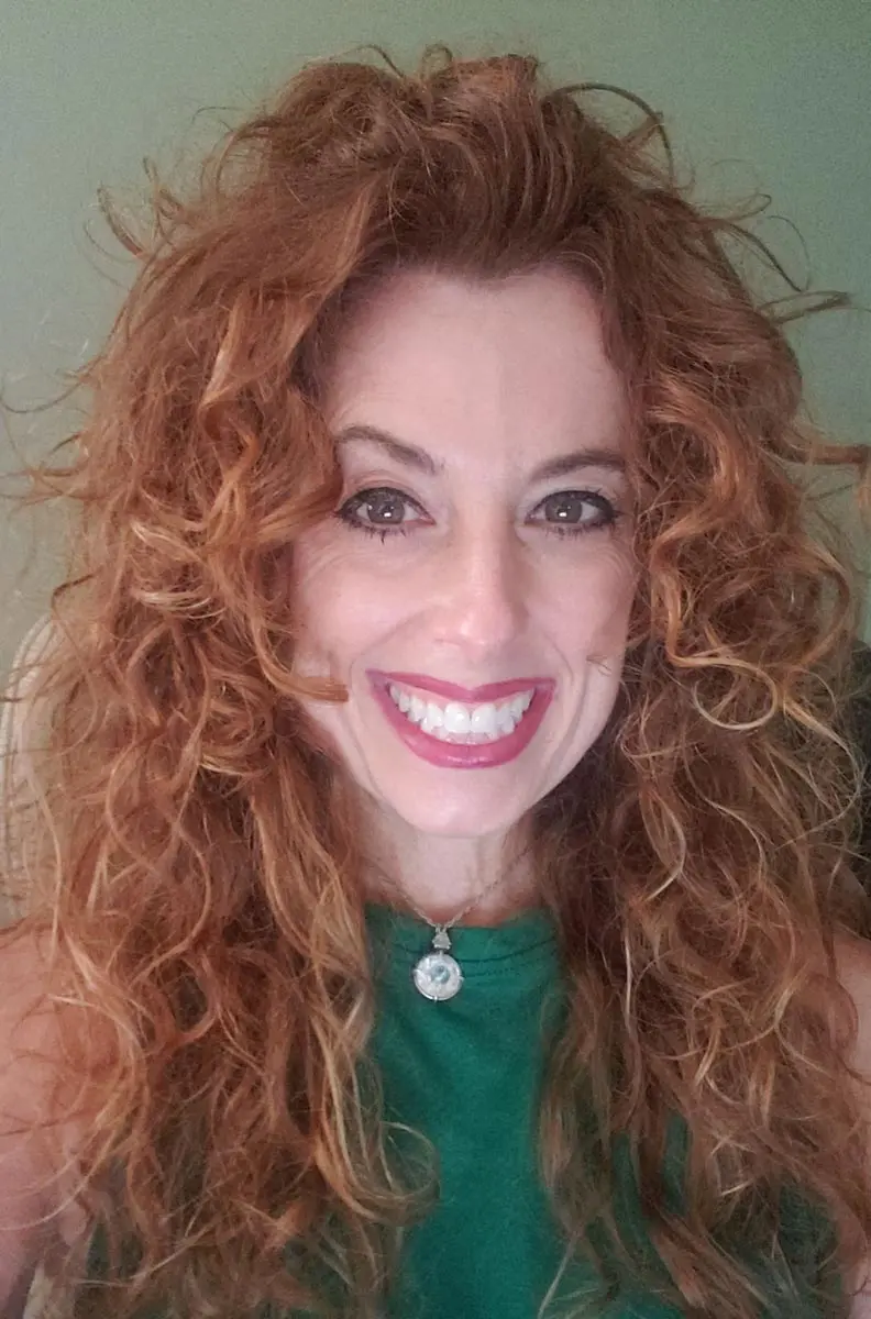 A woman with voluminous curly red hair smiles warmly at the camera, wearing a green top and a necklace.