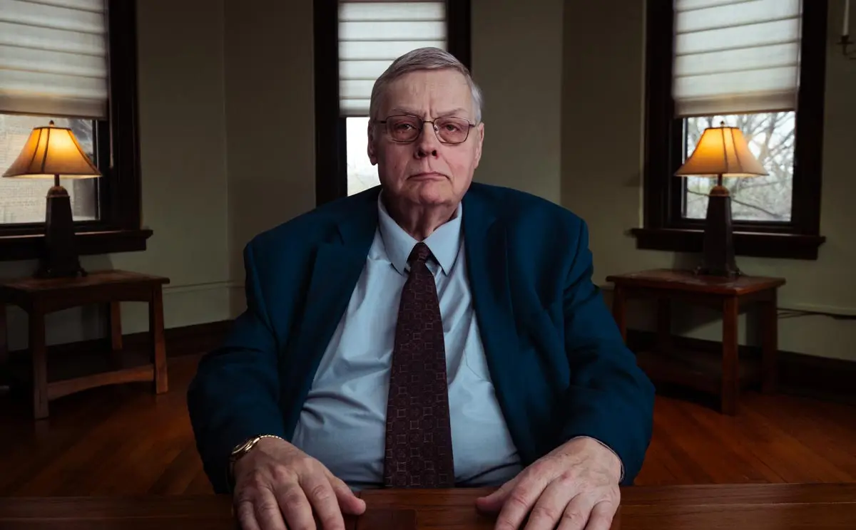 A serious older man in a blue suit sits at a table in a well-lit room with lamps, conveying a sense of authority.