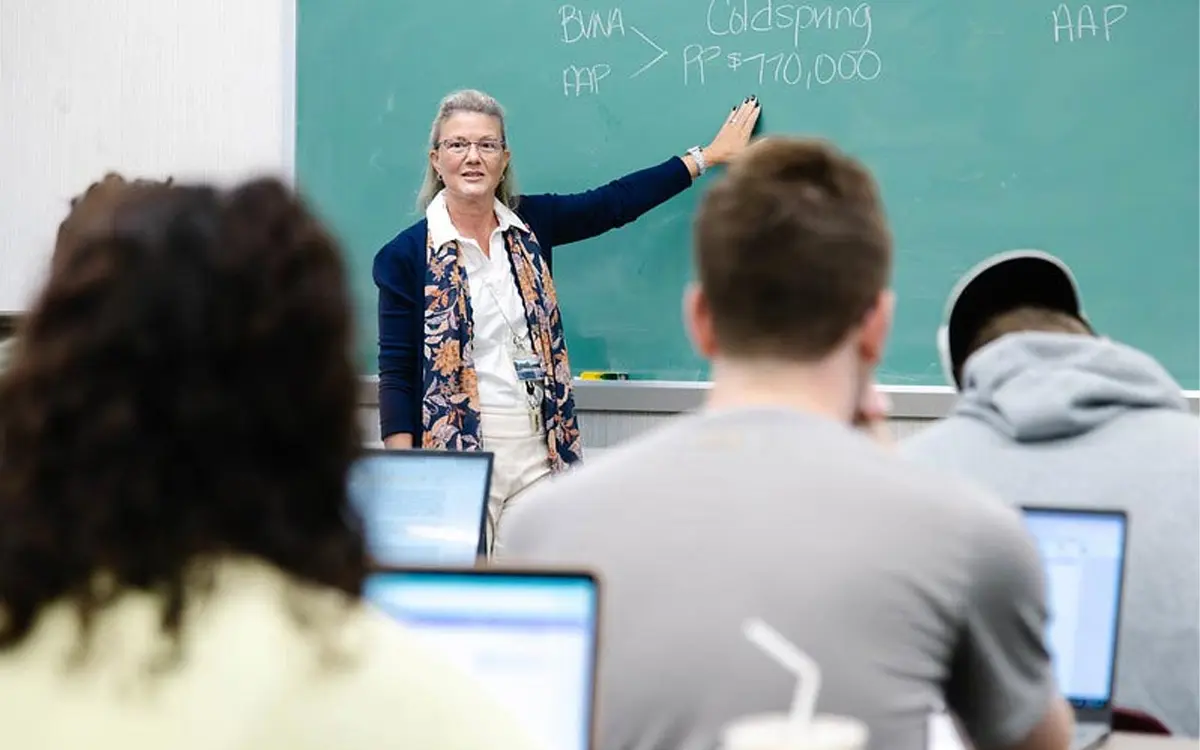 A teacher demonstrates a concept on a chalkboard in a classroom filled with students focused on their laptops.