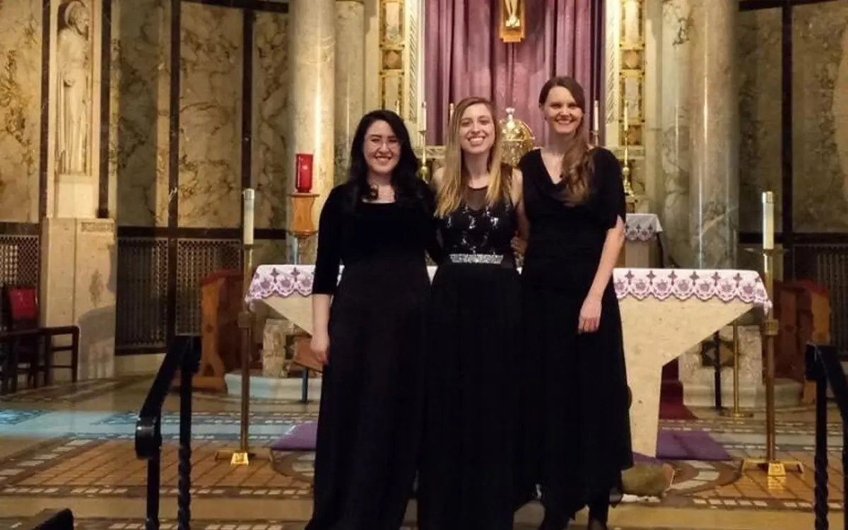 Three women in elegant black dresses pose together in a church setting, with a decorated altar in the background.