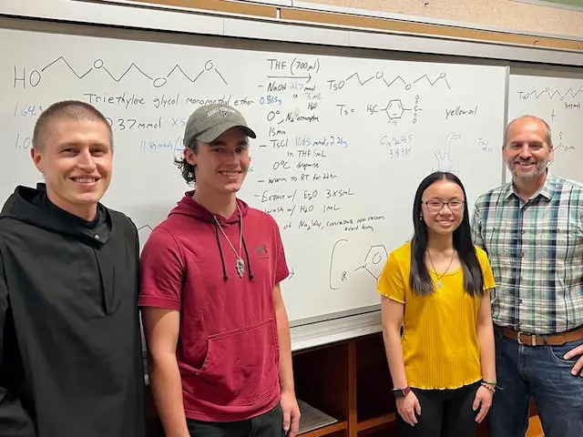 A group of four students stands in front of a whiteboard filled with chemical structures and formulas, showcasing a collaborative learning environment.