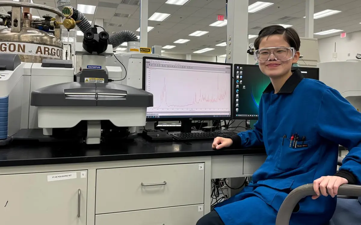 A young scientist in lab attire poses next to a desktop computer displaying scientific data in a laboratory setting.