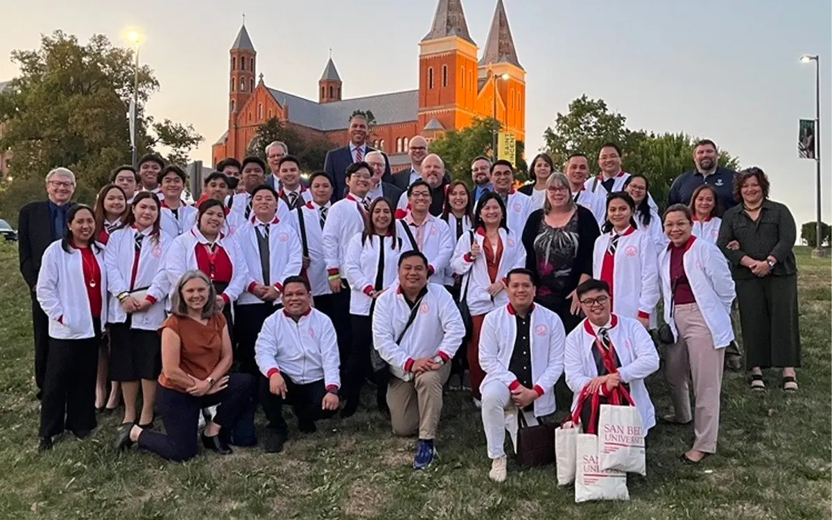 A group photo of a diverse cohort of students and faculty members, all wearing white coats, posing outdoors in front of a historic building at sunset.