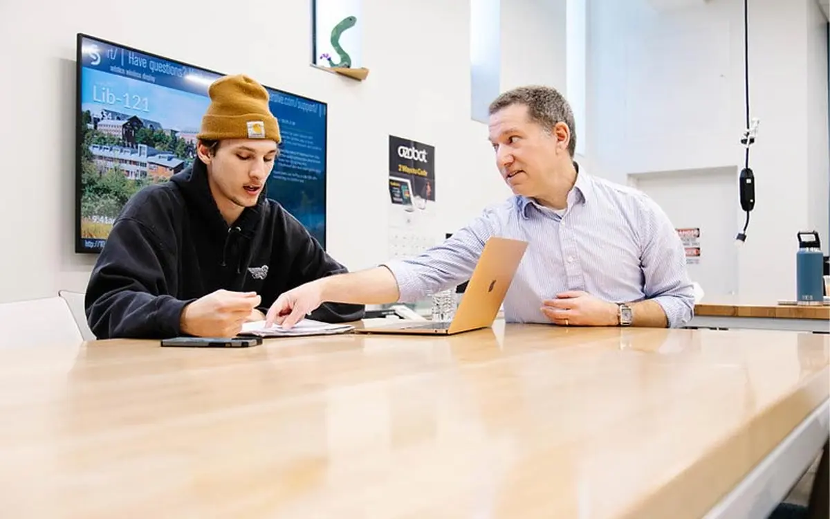 A student and instructor are engaged in a discussion at a table, with a laptop and notes in front of them, highlighting a collaborative learning environment.