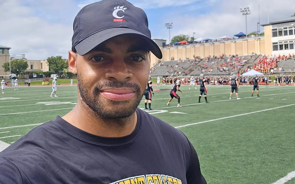 A man wearing a black Cincinnati Bearcats cap poses for a selfie on a football field, with a game in progress and spectators in the background.