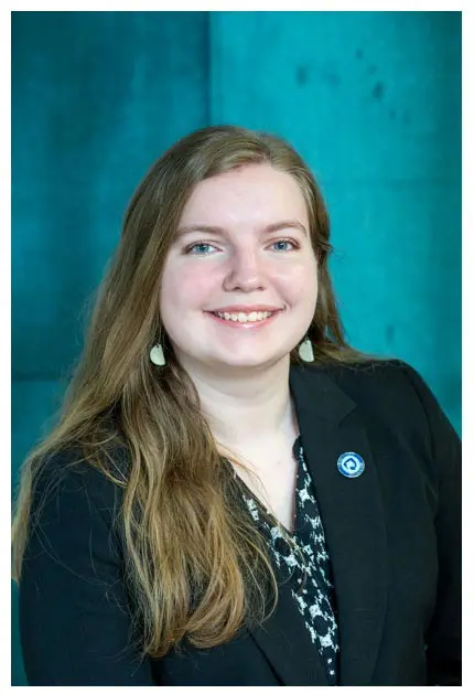 A headshot of a smiling woman with long hair wearing a black blazer and earrings, against a teal background.