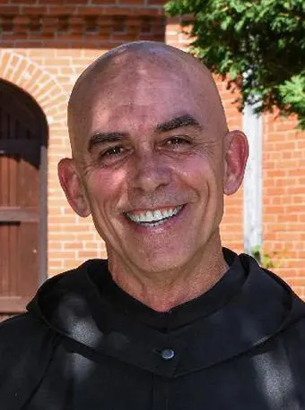 Smiling man in a black robe stands in front of a brick wall and greenery, showcasing a warm and welcoming expression.