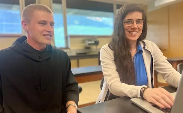 Two professors engaged in a collaborative activity at a classroom table, smiling while working on a laptop.