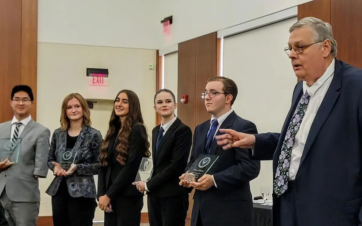 A group of six individuals, dressed in formal attire, stand together while receiving awards at a ceremony, with one person speaking to the audience.