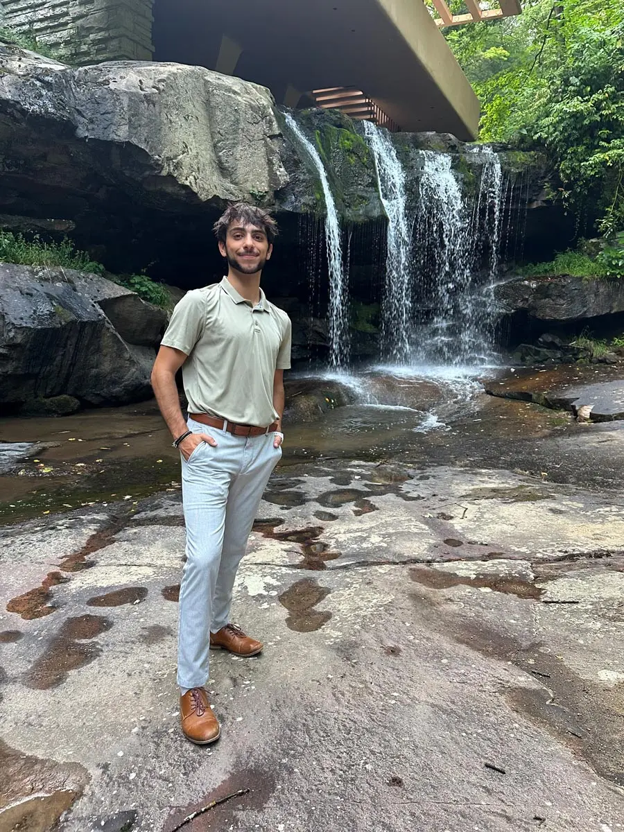 A man standing confidently by a small waterfall and rocky terrain, dressed in casual attire.