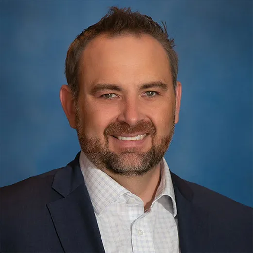 Professional headshot of a man with a beard, wearing a suit jacket and collared shirt, set against a blue background.