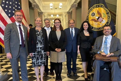 A group of seven professionals poses for a photo in a formal setting, surrounded by the U.S. flag and an official seal.