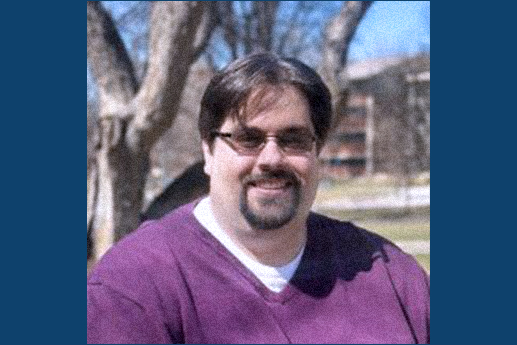 Smiling man in a purple shirt sitting outdoors with trees in the background.