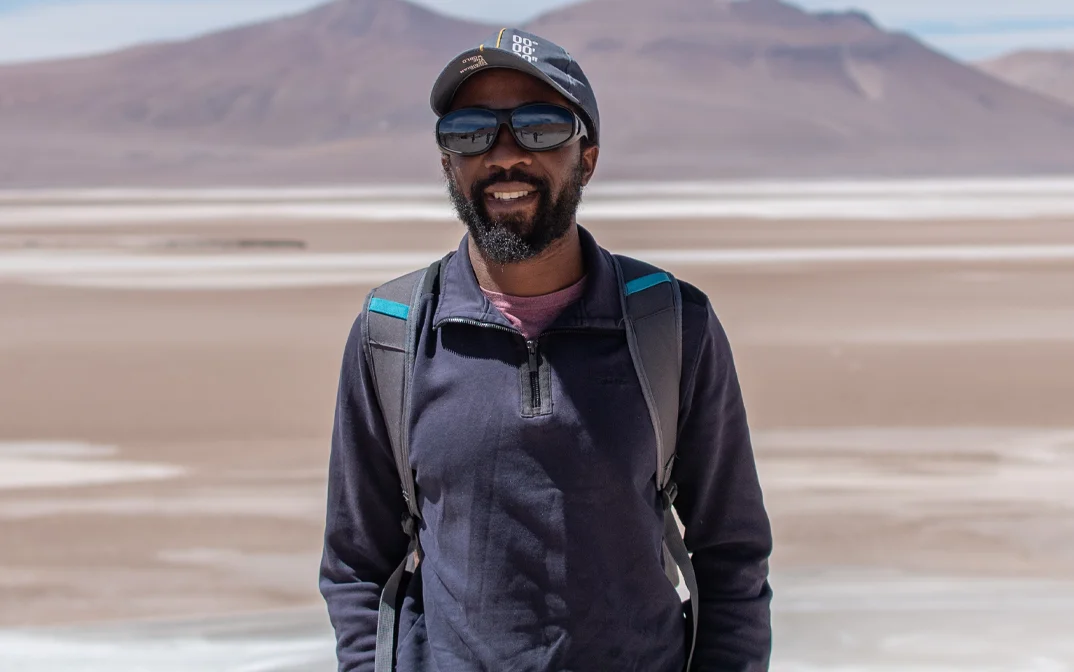 A smiling man with a backpack stands in front of a vast salt flat and distant mountains.