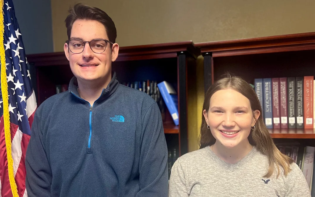 Two smiling students standing in front of a bookshelf, with an American flag to the left.