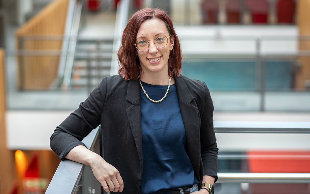 A professional woman with red hair and glasses poses confidently on a balcony in a modern building.