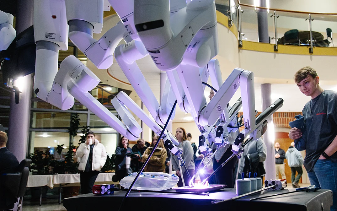 A modern robotic system is showcased at an event, with multiple robotic arms in action and onlookers observing the demonstration.