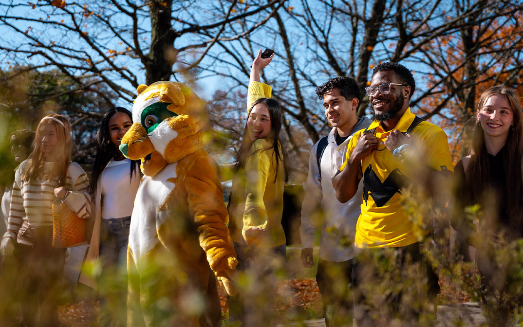 A group of enthusiastic students dressed in yellow, including a mascot in a cheerful costume, celebrating outdoors among autumn foliage.