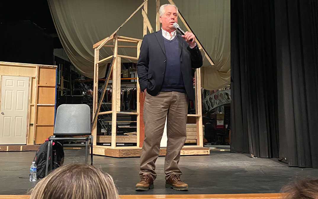 a man stands on a stage holding a microphone addressing students in the audience.