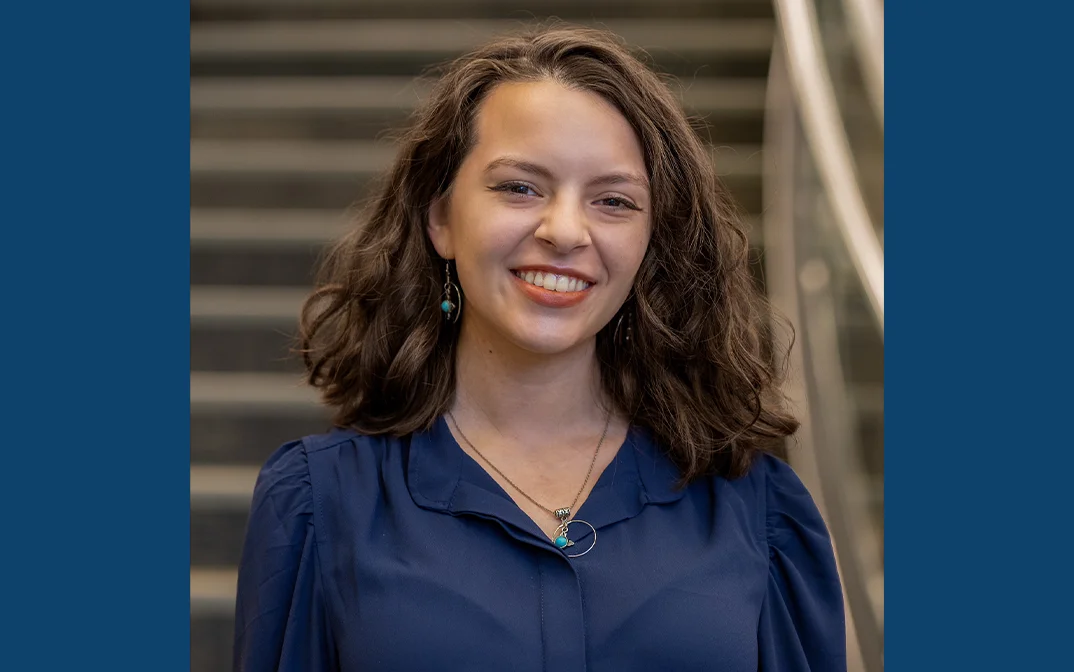 A smiling young woman with curly hair wearing a blue blouse, standing in front of a staircase.