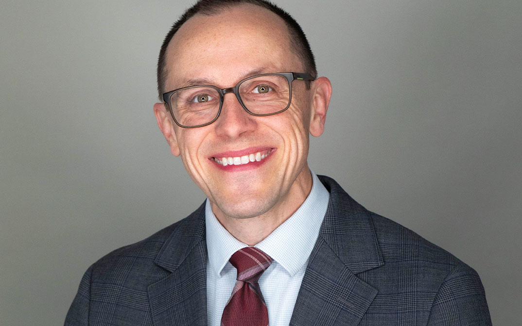 Professional headshot of a man in a suit with glasses, smiling confidently at the camera.