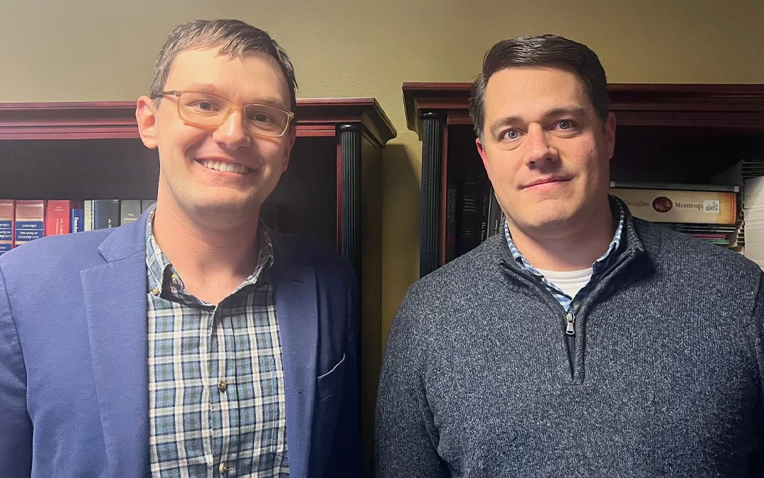 Two men are smiling for the camera in an office setting, standing beside bookshelves filled with legal books and documents.