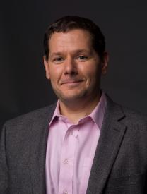 Professional headshot of a man in a gray blazer and light pink shirt.