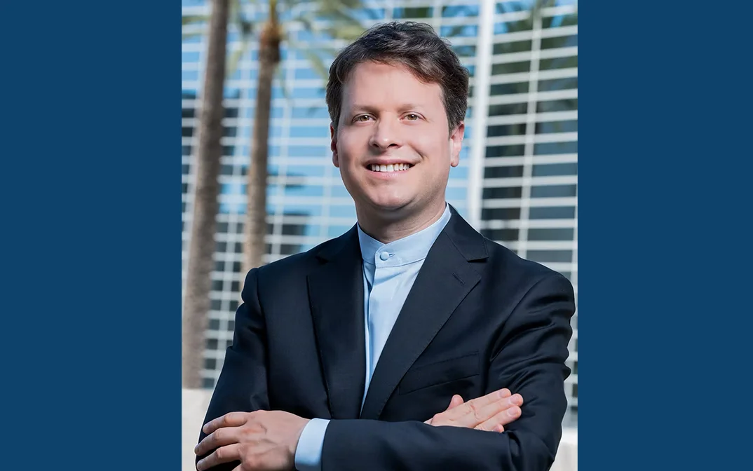 Portrait of a smiling man in a suit with crossed arms, standing outdoors in front of a modern building and palm trees.